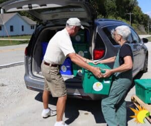 A man and a woman work together to unload crates of food from the back of a van.